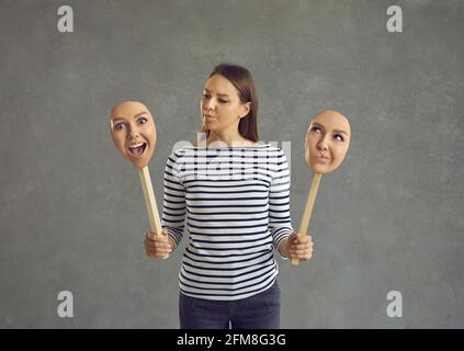 Frau steht auf einem grauen Hintergrund und hält zwei Gesichter mit unterschiedlichen Emotionen und Stimmungen. Stockfoto