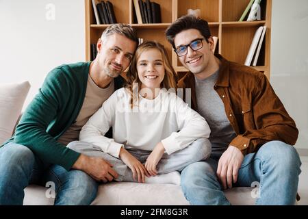 Fröhliche, schwule Familie, die lächelt und die Kamera anschaut, während sie sitzt Auf der Couch zu Hause Stockfoto