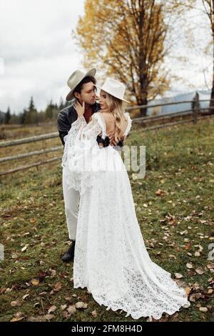 Bezauberndes Modepaar, das auf dem Feld steht, umarmt und ein Wochenende zusammen verbringt. Charming Mann mit Blick auf schöne stilvolle Frau in weißem Kleid Stockfoto