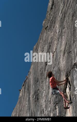 Ein junger Mann in einem roten T-Shirt und blauen Shorts klettert auf einen grauen Felsen mit einem blauen Himmel im Hintergrund. Stockfoto