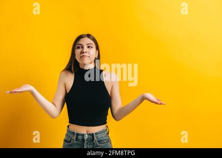 Junge kaukasische Frau mit brünetten Haaren zuckende Missverständnisse, Porträt der emotionalen Frau isoliert auf gelbem Hintergrund mit Copy Space. Stockfoto