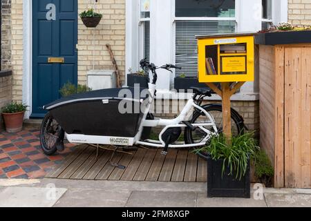 Kleine freie Bibliothek neben einem elektrischen Kind von Urban Arrow, das Fahrrad trägt. Cambridge, Großbritannien. Stockfoto