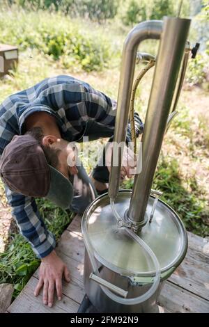 Fokussierter Mann macht an einem Sommertag in einem Dorfhof eine Alkoholmashine. Zubereitung eines alkoholischen Getränks. Stockfoto