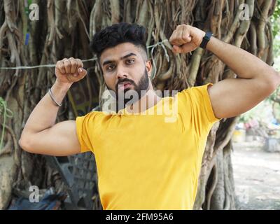 Porträt eines hübschen Indianers in einem gelben T-Shirt Zeigt diesen Bizeps vor einem Baumhintergrund Stockfoto