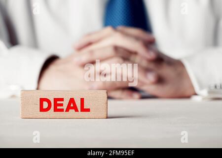 Das Wort Deal auf einem Holzblock vor einem Geschäftsmann mit gekreuzten Händen geschrieben. Business Deal-Konzept Stockfoto