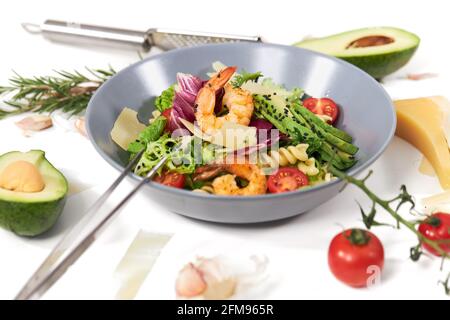 Nahaufnahme eines modernen blauen Tellers mit Gemüsesalat mit Pasta, Avocado und Garnelen auf weißem Hintergrund. Konzept der Prozessküche gesunde und appetitliche Salat zu Hause. Stockfoto