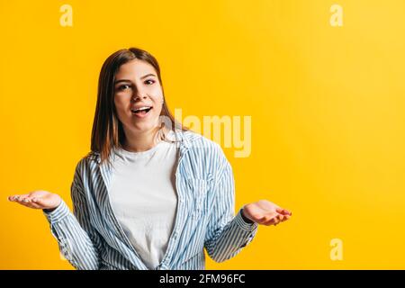 Junge kaukasische Frau mit brünetten Haaren zuckende Missverständnisse, Porträt der emotionalen Frau isoliert auf gelbem Hintergrund mit Copy Space. Stockfoto