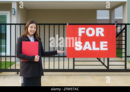Die asiatische Frauenimmobilienagentur steht vor der Tür Das Haus mit Verkaufsschild hängt vor der Haustür Geben Sie bekannt, dass sich Interessierte in CO befinden Stockfoto