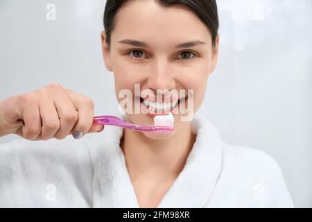 Vorderansicht Porträt einer lächelnden jungen Brünette Frau mit rosa Zahnbürste und Zahnpasta im Badezimmer. Konzept des Prozesses Vorbereitung auf die Reinigung der Zähne mit guter Laune. Stockfoto