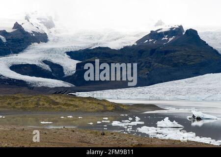 Eis vom Breioamerkurjokull-Gletscher (hinten) kält sich in den Fjallsarlon-Gletschersee im Vatnajokull-Nationalpark, Island. Stockfoto