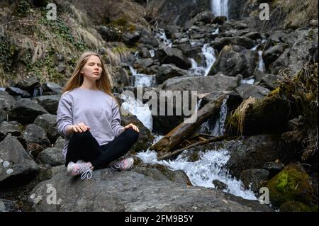 Mädchen sitzt auf einem großen Stein und meditiert in der Nähe eines Wasserfalls neben einem Bergfluss. Stockfoto