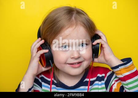 Fröhliches, lächelndes kleines Kind, das Musik hört, mit großen Kopfhörern, isoliert auf gelbem Hintergrund. Stockfoto