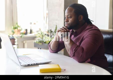 Fokussierter und besorgter schwarzer Mann in elegantem Casual-Shirt sieht auf den Laptop-Bildschirm, seriös afroamerikanischer Kerl sitzt am Schreibtisch, das Kinn auf den Händen ruht, fühlt Zweifel und löst schwierige Aufgaben Stockfoto