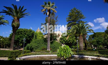 Italien, Sizilien, Südost-Sizilien, Barockwinkel, Ragusa, Am späten Morgen, blauer Himmel mit Wolken, Giardino Ibleo Park, Brunnen, dahinter drei Palmen, Kirche Chiesa di San Vincenzo Ferreri im Hintergrund hinter Büschen versteckt Stockfoto