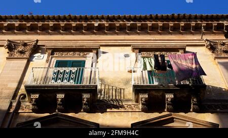 Italien, Sizilien, Südost-Sizilien, Barockwinkel, Ragusa, Morgenlicht, Altsatdt, Blick von schräg unten auf zwei Balkone eines Barockhauses, über einem Streifen blauen Himmels, Wäsche hängt auf dem rechten Balkon, grüne Türfensterläden Stockfoto