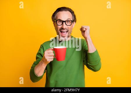 Fotoportrait eines Mannes in einer Brille, der wie ein Gewinner beim Trinken gestikuliert Kaffee in der Pause isoliert auf lebhaft gelbem Hintergrund Stockfoto