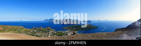 Italien, Sizilien, Äolische Inseln, Panoramablick, Vulcano, Krater, Blick vom Krater auf sechs Äolische Inseln (von links nach rechts): Alicudi, Filicudi, Salina, Lipari, Panarea, Stromboli, blauer Himmel. Schiff fährt in den Hafen von Vulcano ein Stockfoto