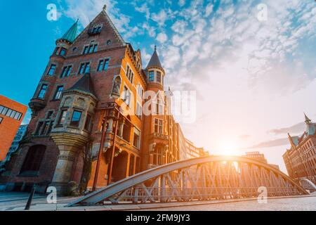 Metallbogenbrücke und altes rotes Ziegelgebäude in der Speicherstadt der Hamburger HafenCity mit Sonnenblitzen bei Sonnenuntergang, goldener Stunde und weißen Wolken am blauen Himmel darüber. Stockfoto