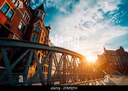 Metallbogenbrücke und altes rotes Ziegelgebäude in der Speicherstadt der Hamburger HafenCity mit Sonnenblitzen bei Sonnenuntergang, goldener Stunde und weißen Wolken am blauen Himmel darüber. Stockfoto