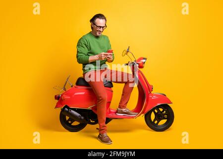 Foto von Mann halten Telefon Fahrt Roller tragen Brille grün Pullover Hose Schuhe isoliert gelben Hintergrund Stockfoto