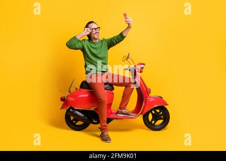 Foto von Kerl halten Smartphone Fahrt Moped nehmen Selfie tragen Specs grün Pullover Hose Schuhe isoliert gelbe Farbe Hintergrund Stockfoto