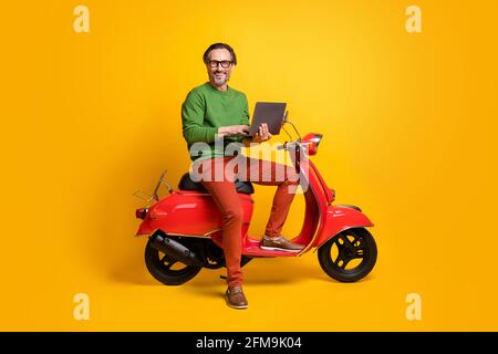 Foto von Mann halten Laptop Fahrt Roller tippen Tragen Sie eine Brille Grün Pullover Hose Schuhe isoliert gelbe Farbe Hintergrund Stockfoto