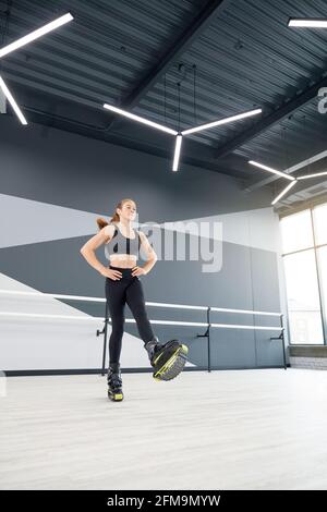 Junge kaukasische Mädchen mit Pferdeschwanz üben kangoo springen drinnen. Von unten sehen Sie, wie eine Sportlerin die Hände an der Taille hält und Cardio-Dance-Workout in der Halle genießt, Hi-Tech-Innenausstattung. Stockfoto