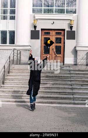 Glückliche junge Frau in schwarzem Abschlusskleid und schützendem Gesicht Maske wirft Graduate Cap mit gelben Quaste auf dem Hintergrund Der Universität oder Hochschule e Stockfoto
