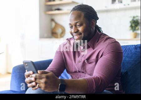 Lächelnder afroamerikanischer Mann mit Smartphone sitzt zu Hause auf dem Sofa, verbringt seine Freizeit mit Videospielen, scrollt in den Neuigkeiten in den sozialen Medien, chattet online und nutzt mobile Apps Stockfoto