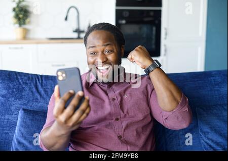Ihr habt Glück. Urkomischer afroamerikanischer Mann mit Smartphone, der zu Hause auf dem Sofa sitzt, den Sieg feiert, in der Online-Lotterie gewinnt, glücklich mit dem Sieg des Sportteams Stockfoto
