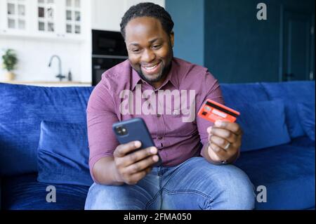 Afroamerikanischer Mann, der online bezahlt, Transaktionen auf seinem Bankkonto macht, Smartphone und Kreditkarte hält, online einkauft und zu Hause auf der Couch Essen bestellt Stockfoto