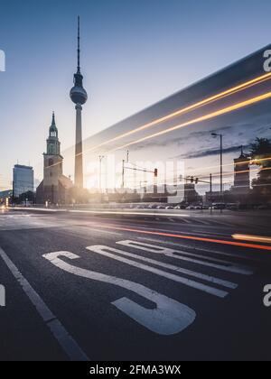 Leichte Spur des Straßenverkehrs auf dem Alexanderplatz mit dem Berliner Fernsehturm und der Marienkirche bei Sonnenaufgang. Stockfoto