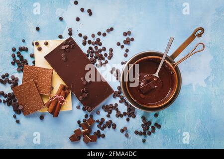 Geschmolzene Schokolade in Kupferpfanne und Tafeln aus dunkler und weißer Schokolade daneben auf rustikaler blauer Oberfläche. Draufsicht, Leerraum Stockfoto