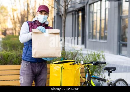 Junger Kurier in medizinischer Maske liefert Lebensmittel mit gelbem Thermalrucksack, fährt mit dem Fahrrad in der Stadt. Konzept für den Lieferservice von Lebensmitteln Stockfoto