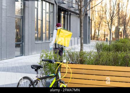 Junger Kurier in medizinischer Maske liefert Lebensmittel mit gelbem Thermalrucksack, fährt mit dem Fahrrad in der Stadt. Konzept für den Lieferservice von Lebensmitteln Stockfoto