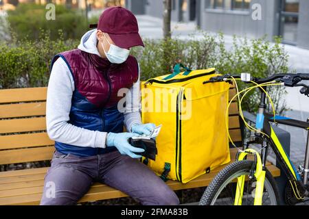 Junger Kurier in medizinischer Maske liefert Lebensmittel mit gelbem Thermalrucksack, fährt mit dem Fahrrad in der Stadt. Konzept für den Lieferservice von Lebensmitteln Stockfoto