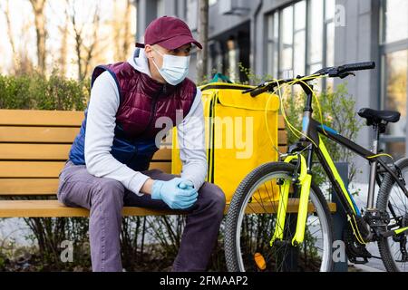 Junger Kurier in medizinischer Maske liefert Lebensmittel mit gelbem Thermalrucksack, fährt mit dem Fahrrad in der Stadt. Konzept für den Lieferservice von Lebensmitteln Stockfoto