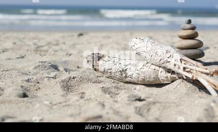 Getrockneter weißer Salbei-Rauch, Verwischungsstäbchen brennend im weichen Fokus mit Bokeh, Aroma verschmiert aus nächster Nähe. Felsbalancierung am Meeresstrand, Steine stapeln sich von Meereswellen. Zen-Pyramide aus Kieselsteinen am Sandstrand Stockfoto