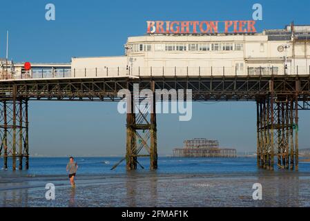 Der verfallene West Pier von Brighton wurde in die Struktur des „Brighton Pier“, des Palace Pier, eingefasst. Mann beim Joggen am Ufer im Vordergrund. Stockfoto