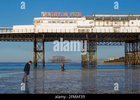 Der verfallene West Pier von Brighton wurde in die Struktur des „Brighton Pier“, des Palace Pier, eingefasst. Fischer sammeln Ragworm für Köder im Vordergrund, Frauen gehen für ein Schwimmen hinter. Stockfoto