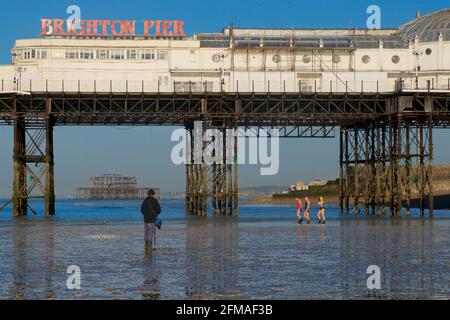 Der verfallene West Pier von Brighton wurde in die Struktur des „Brighton Pier“, des Palace Pier, eingefasst. Fischer sammeln Ragworm für Köder im Vordergrund, Frauen gehen für ein Schwimmen hinter. Stockfoto