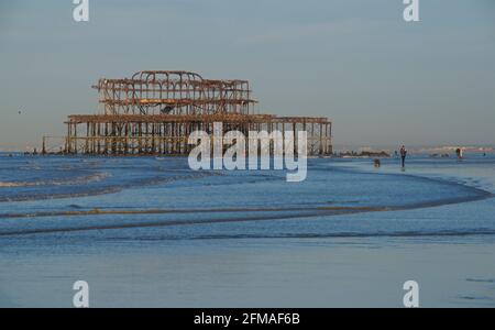 Menschen, die bei Ebbe am frühen Morgen am Strand spazieren gehen, mit den rostigen Überresten des heruntergekommenen West Pier von Brighton. Brighton & Hove, Sussex, England, Großbritannien Stockfoto