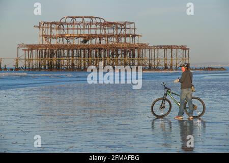 Radler, der bei Ebbe mit dem Mountainbike auf dem Sand steht, Brighton, mit den rostigen Resten des West Pier im Hintergrund. Brighton & Hove, Sussex, England