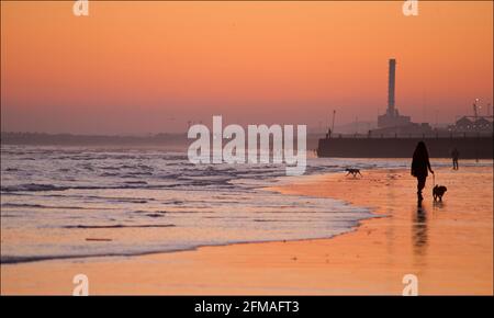 Brighton und Hove Beach bei Ebbe mit Blick nach Westen in Richtung Shoreham Power Station. Silhouetten von Menschen, die bei Sonnenuntergang am Sandstrand entlang wandern. East Sussex, England. Gehen Sie mit dem Hund. Stockfoto