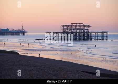 Bei Ebbe schlendern die Menschen am frühen Morgen am Strand entlang, wobei die rostigen Überreste von Brighton's verfallenen West Pier und dem Palace Pier dahinter vor dem rosafarbenen Glanz des Himmels silhouettiert wurden. Brighton & Hove, Sussex, England, Großbritannien Stockfoto