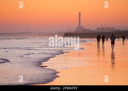 Brighton und Hove Beach bei Ebbe mit Blick nach Westen in Richtung Shoreham Power Station. Silhouetten von Menschen, die bei Sonnenuntergang am Sandstrand entlang wandern. East Sussex, England Stockfoto