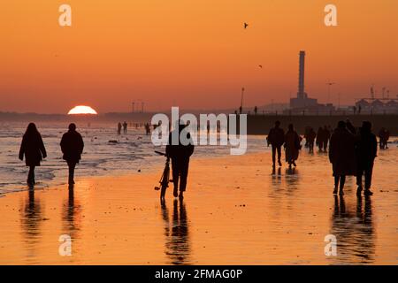 Brighton und Hove Beach bei Ebbe mit Blick nach Westen in Richtung Shoreham Power Station. Silhouetten von Menschen, die bei Sonnenuntergang am Sandstrand entlang wandern. East Sussex, England Stockfoto