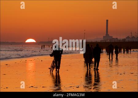 Brighton und Hove Beach bei Ebbe mit Blick nach Westen in Richtung Shoreham Power Station. Silhouetten von Menschen, die bei Sonnenuntergang am Sandstrand entlang wandern. East Sussex, England Stockfoto