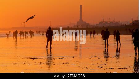 Brighton und Hove Beach bei Ebbe mit Blick nach Westen in Richtung Shoreham Power Station. Silhouetten von Menschen, die bei Sonnenuntergang am Sandstrand entlang wandern. East Sussex, England Stockfoto