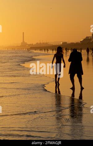 Brighton und Hove Beach bei Ebbe und Blick nach Westen. Silhouetten von Menschen, die bei Sonnenuntergang am Sandstrand entlang wandern. East Sussex, England. Mann, der eine Frau gerade aus dem Schwimmen fotografiert. Stockfoto